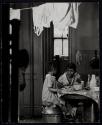 [Little girl seated on a milk can at table, with woman]
