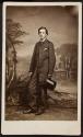 [Young man standing with top hat and umbrella in front of scenic backdrop]