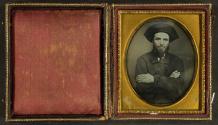 [Man in wide-brimmed hat (folded back), overcoat, arms folded across chest]