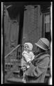 [Woman with cloche hat, veil, holding baby in front of brownstone]