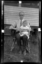 [Man holding baby, seated on chair, outdoors]