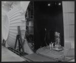 [Two technicians working on a satellite in a testing bay.]