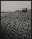 [A worker inspects rolls of metal wire lined up outside a factory.]