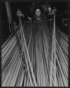 [Worker lifting two pieces of thick wire from a large pile in front of her.]