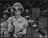 [Woman  in factory holding canister looking through attached curled pipes]