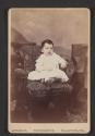 [Small boy sitting in floral chair wearing a white gown and a locket]