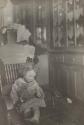 [Child seated in rocking chair, holding a top and a candle]
