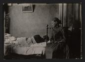 [Child in chair overlooking adult in bed in tenement bedroom]