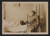 [Child in chair overlooking adult in bed in tenement bedroom]