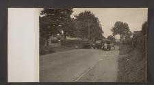 [A woman buys food from a grocery truck on a town road as two trucks pass pass by.]