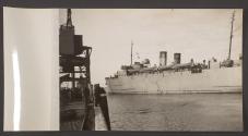 [Troop shipping pulling away from a quay, its railings are lined with people.]