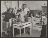 [Teenage girl photographing group of friends baking a cake in a kitchen]
