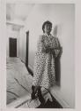 [Woman in flower-print housedress standing in hall]