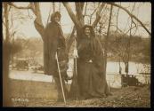 [Two women with cloaks and walking sticks standing in front of trees, members of the Suffrage Hike]