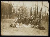 [Group of eleven women seated on lawn with cloaks and walking sticks, banner on the ground]