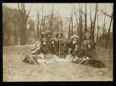 [Group of eleven women seated on the law with cloaks and walking sticks, banner on the ground]