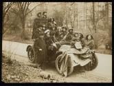 [Group of eleven women, three men and one small girl piled onto an automobile with signs advocating Votes for Women]
