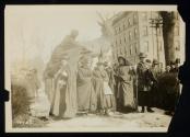 [Six women with cloaks and walking sticks standing in front of sculpture of an elk]