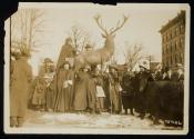 [Eight women with cloaks, walking sticks and votes for women bags, standing in front of a sculpture of an elk with a crowd gathered around]