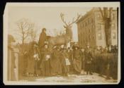 [Seven women with cloaks, walking sticks and votes for women bags, standing in front of a sculpture of an elk with a crowd gathered around]