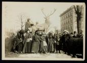 [Eight women with cloaks, walking sticks and votes for women bags, standing in front of a sculpture of an elk with a crowd gathered around]