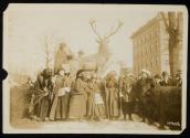 [Eight women with cloaks, walking sticks and votes for women bags, standing in front of a sculpture of an elk with a crowd gathered around]