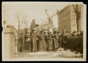 [Seven women with cloaks, walking sticks and votes for women bags, standing in front of a sculpture of an elk with a crowd gathered around]