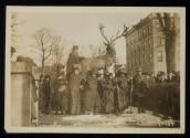 [Seven women with cloaks, walking sticks and votes for women bags, standing in front of a sculpture of an elk with a crowd gathered around]
