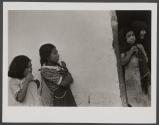 [Two girls beside white wall and girl and older woman inside doorway]
