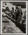 [Women with baskets standing next to river with boats in background]
