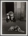 [Man with hat with head down sitting in doorway with dog in foreground]