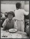 [Child at table with back of woman in background]