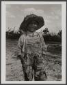 [Portrait of a grinning boy in hat and tattered overalls with cactus in background]