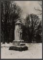 [View of statue of man's head on pedestal with woman and child enveloped into base.]