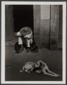[Man with hat with head down sitting in doorway with dog in foreground]